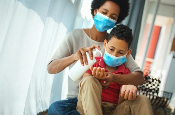 a mother wearing a surgical mask helps her son, also wearing a mask, apply hand sanitizer