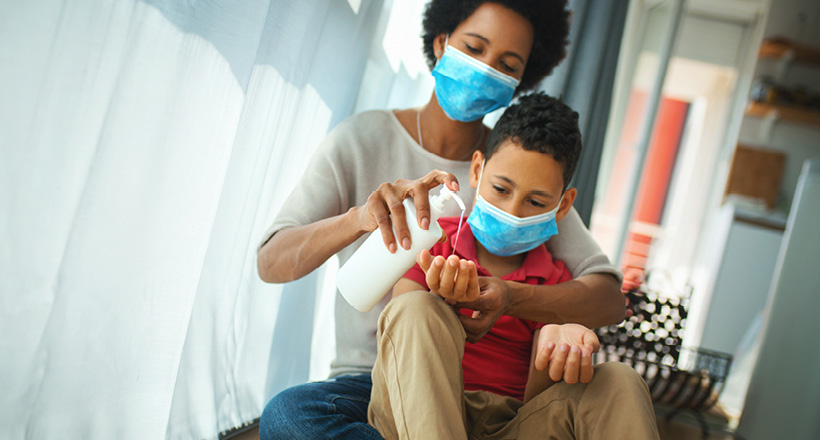 a mother wearing a surgical mask helps her son, also wearing a mask, apply hand sanitizer