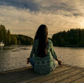 a woman sits in meditation on a deck overlooking a lake and forest. photo courtesy of ANI. 