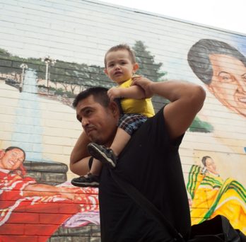 a father carrying his son on his shoulders walking past a mural in the Pilsen neighborhood 