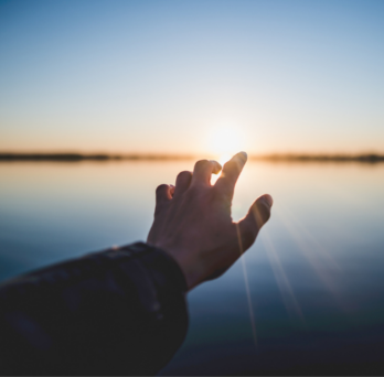 a hand reaches out toward the sun rising over a body of water 