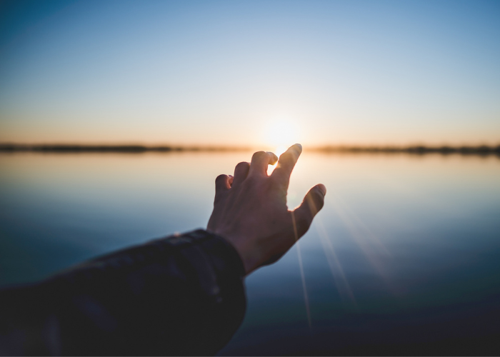 a hand reaches out toward the sun rising over a body of water