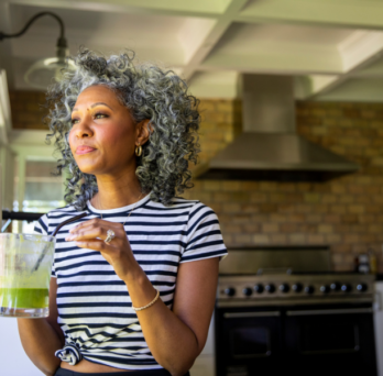 a mature black woman holding a smoothie looks out the window of her kitchen 